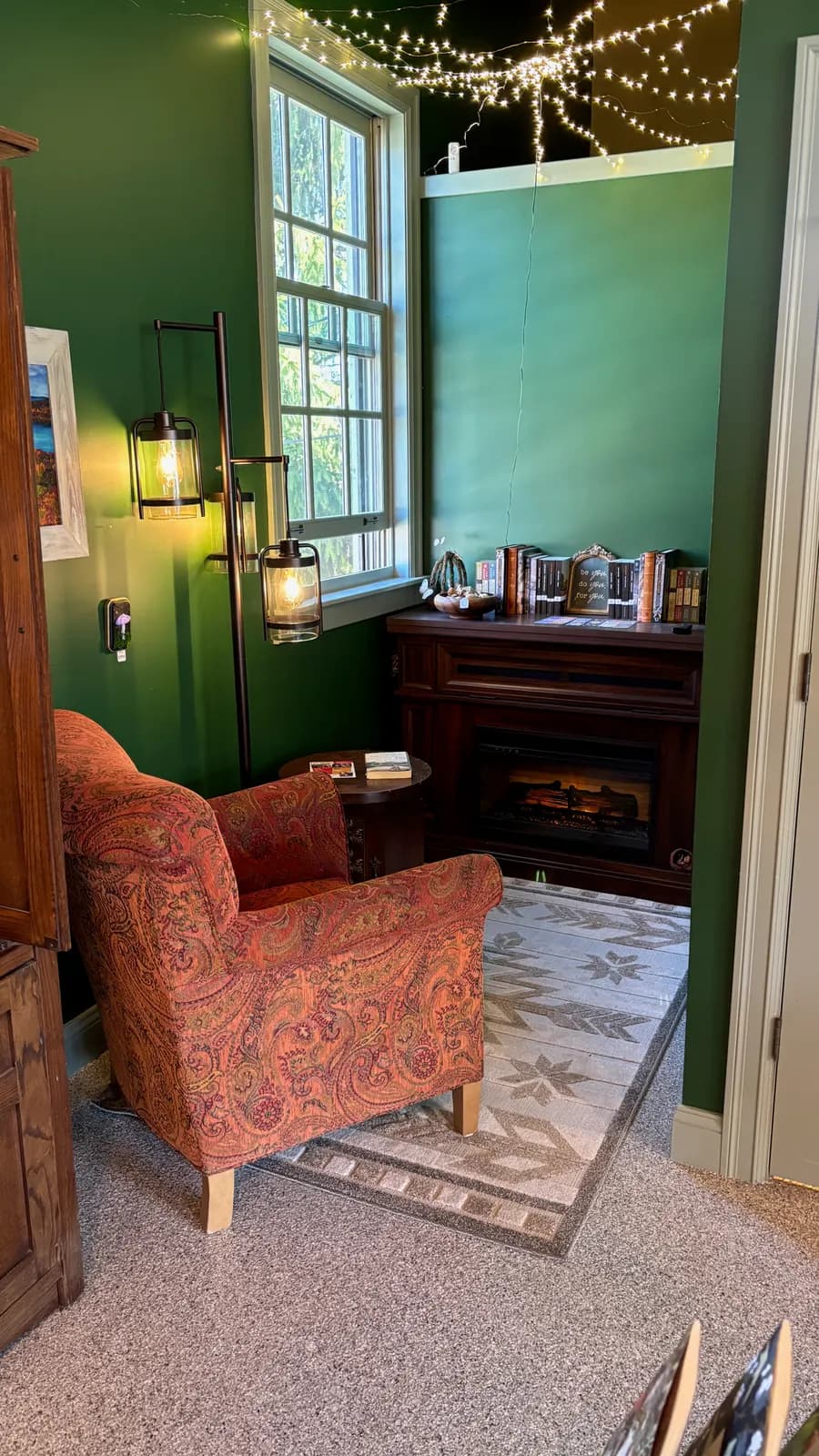 A green sitting room with a worn armchair, a lamp and a small fire